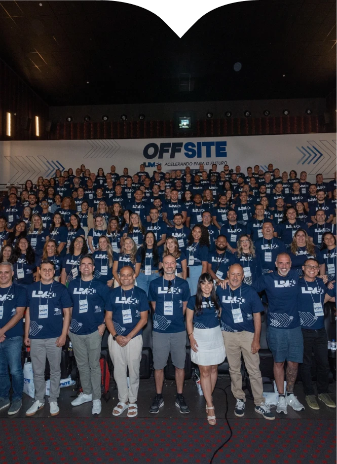 À esquerda da página, um grande grupo de pessoas, entre homens e mulheres, reunidos em um auditório. Vestem camisetas azul-marinho com a inscrição 'L M 50' em branco e crachás de identificação pendurados no pescoço. Ao fundo, uma faixa branca exibe o letreiro centralizado OFFSITE, seguido pela frase com letras menores ACELERANDO PARA O FUTURO, ladeada por grafismos de setas azuis apontando para a direita.