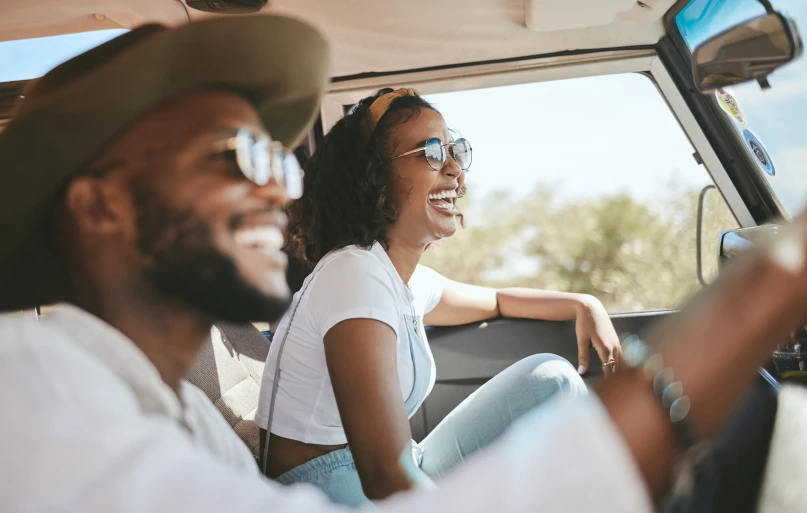 Uma mulher e um homem sorriem dentro de um veículo. A mulher é negra, com cabelos curtos e cacheados. Ela está no assento frontal de passageiro e usa camiseta branca e macacão jeans. Ao lado dela, um homem negro de óculos escuros e barba. Ele está no assento do motorista e usa chapéu bege e camisa branca.